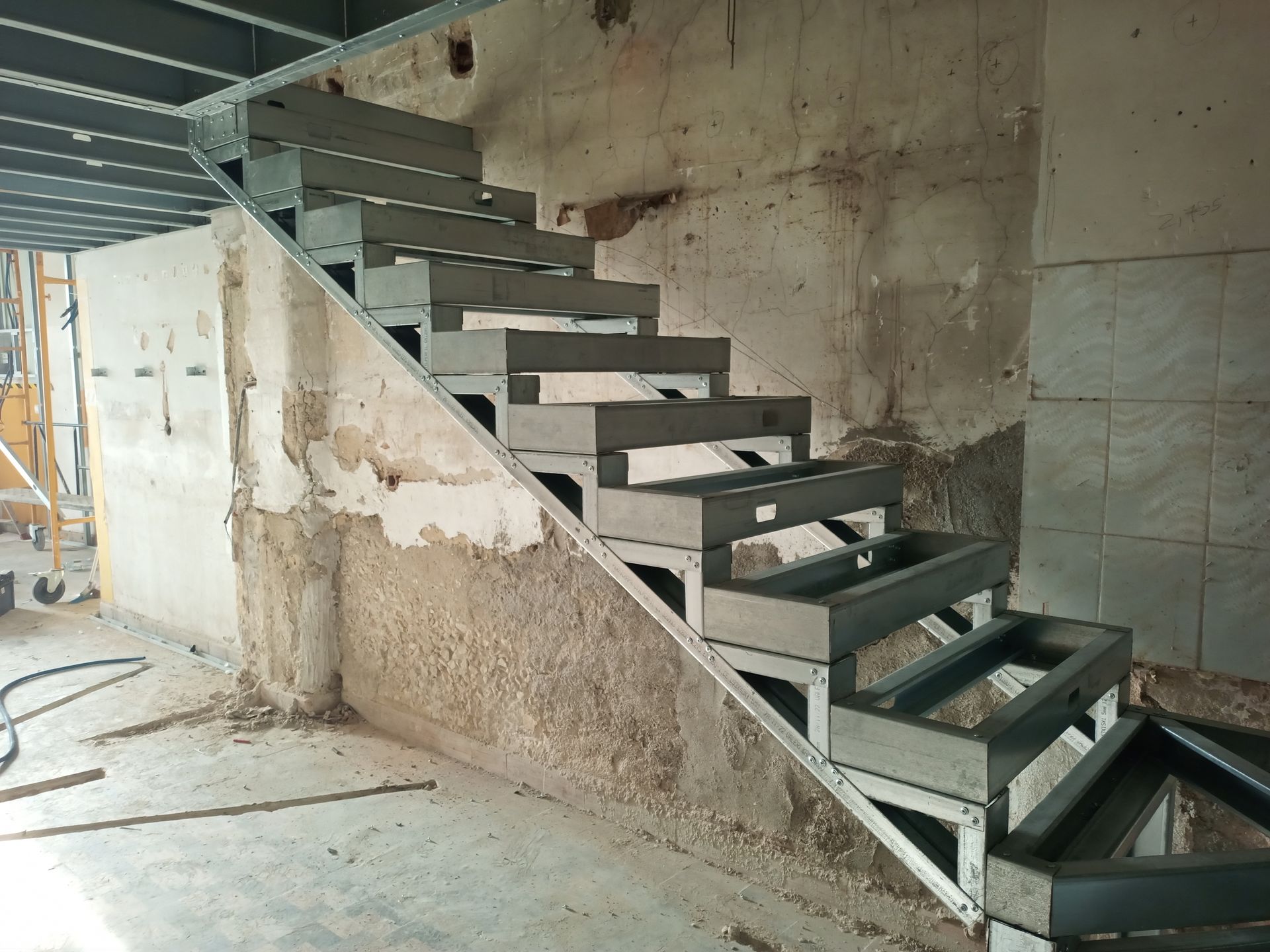 Escada metálica em construção em uma sala com paredes e teto aparentes.