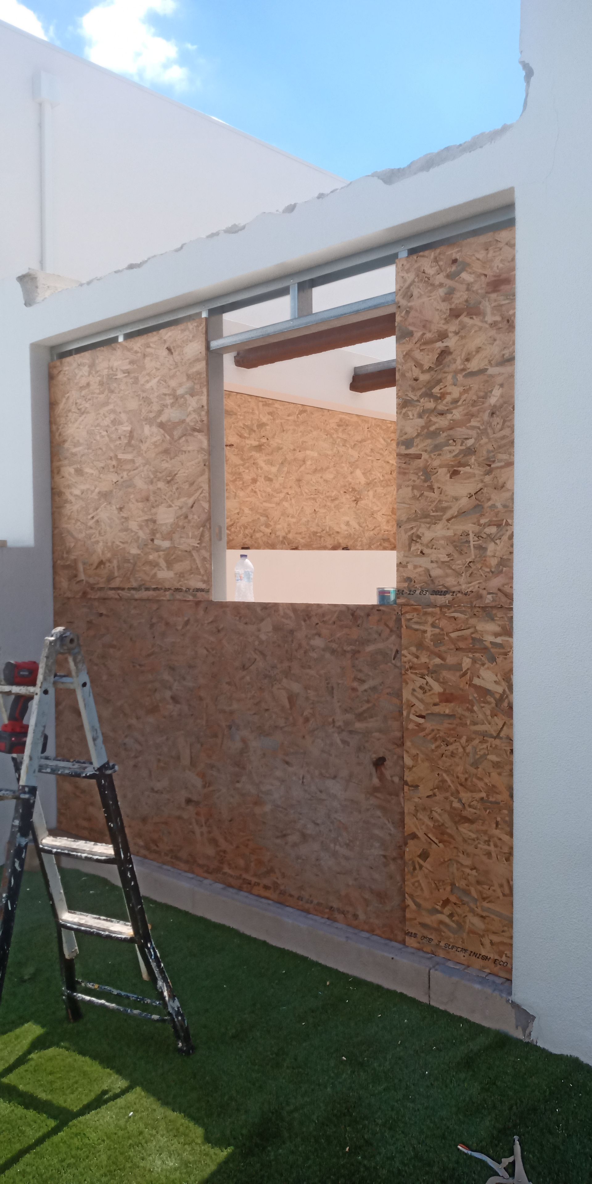 Uma parede com uma janela coberta por placas de madeira compensada, uma escada na grama e céu azul.