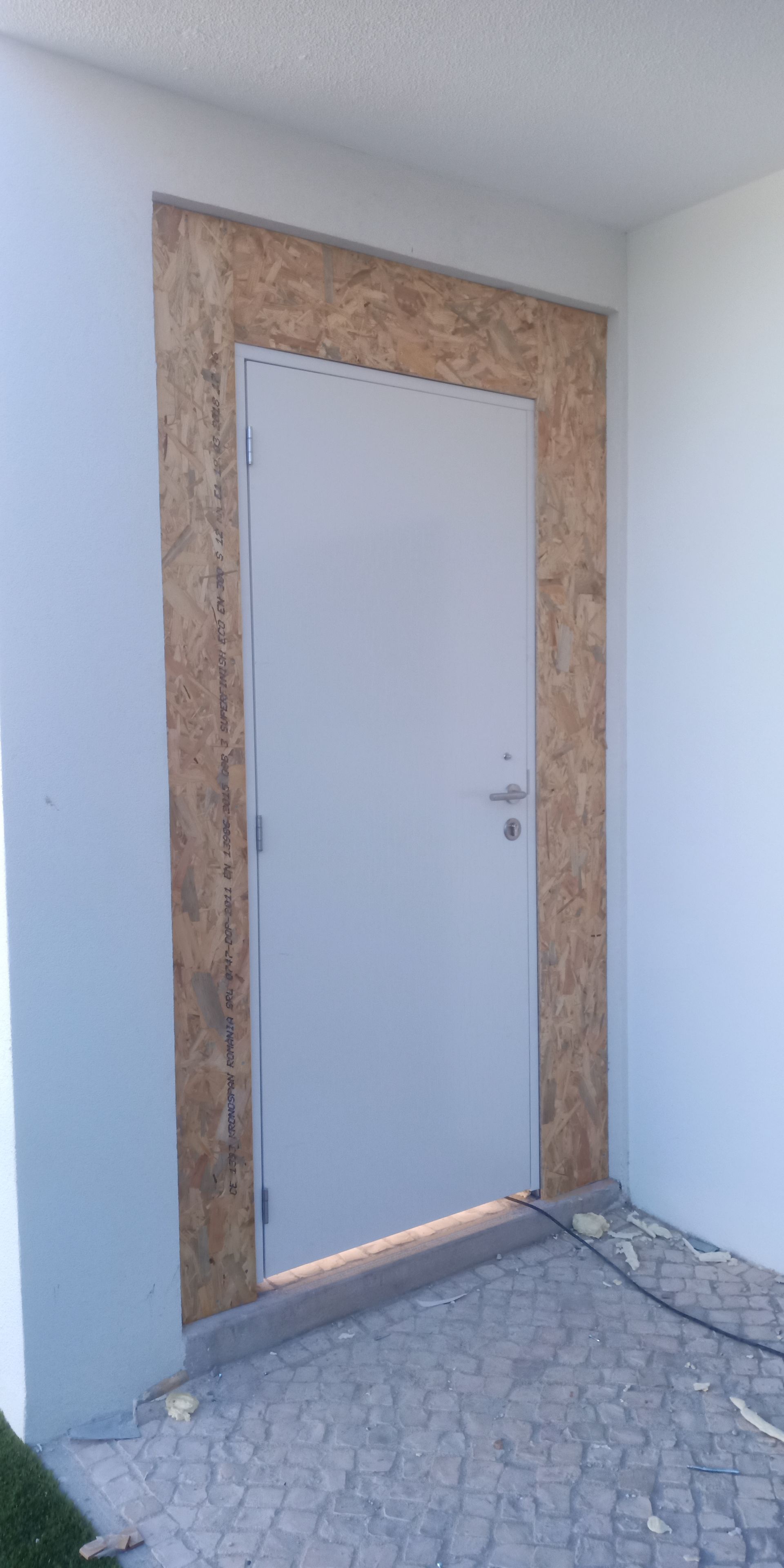 Porta de entrada externa emoldurada com OSB, embutida em parede branca. O piso é formado por lajes de pedra cinza.