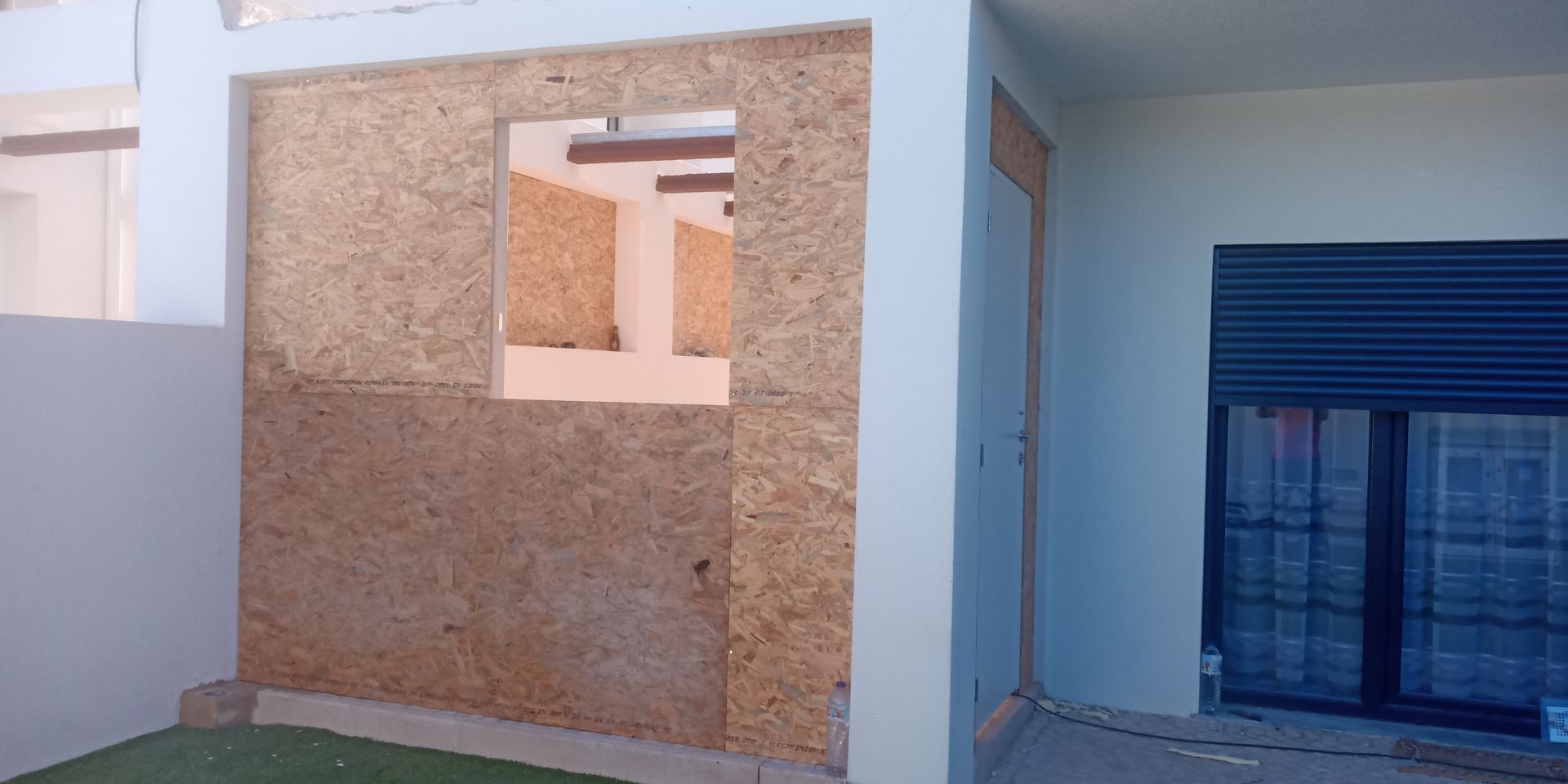 Exterior de um edifício em construção, mostrando uma parede de madeira bruta com uma janela aberta e uma porta fechada.