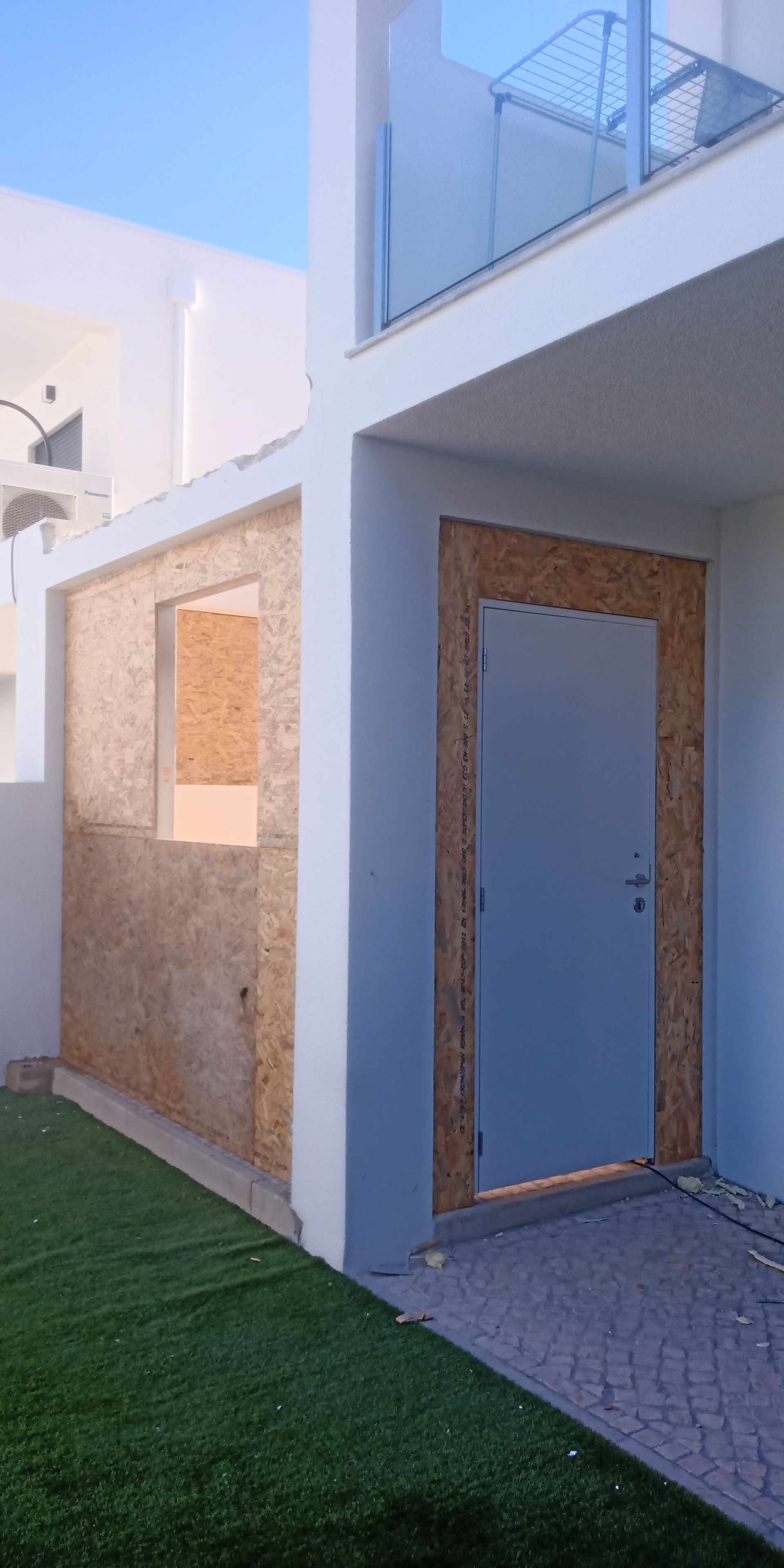 Exterior de um edifício com porta e moldura de janela cinza. A parede externa está inacabada, feita de painéis de madeira.