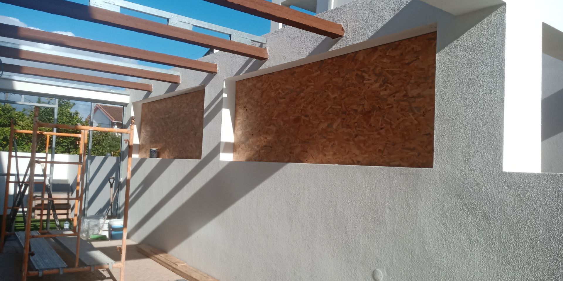 Vista exterior de um edifício com uma parede cinzenta texturizada, vigas de madeira castanhas e céu azul visível.