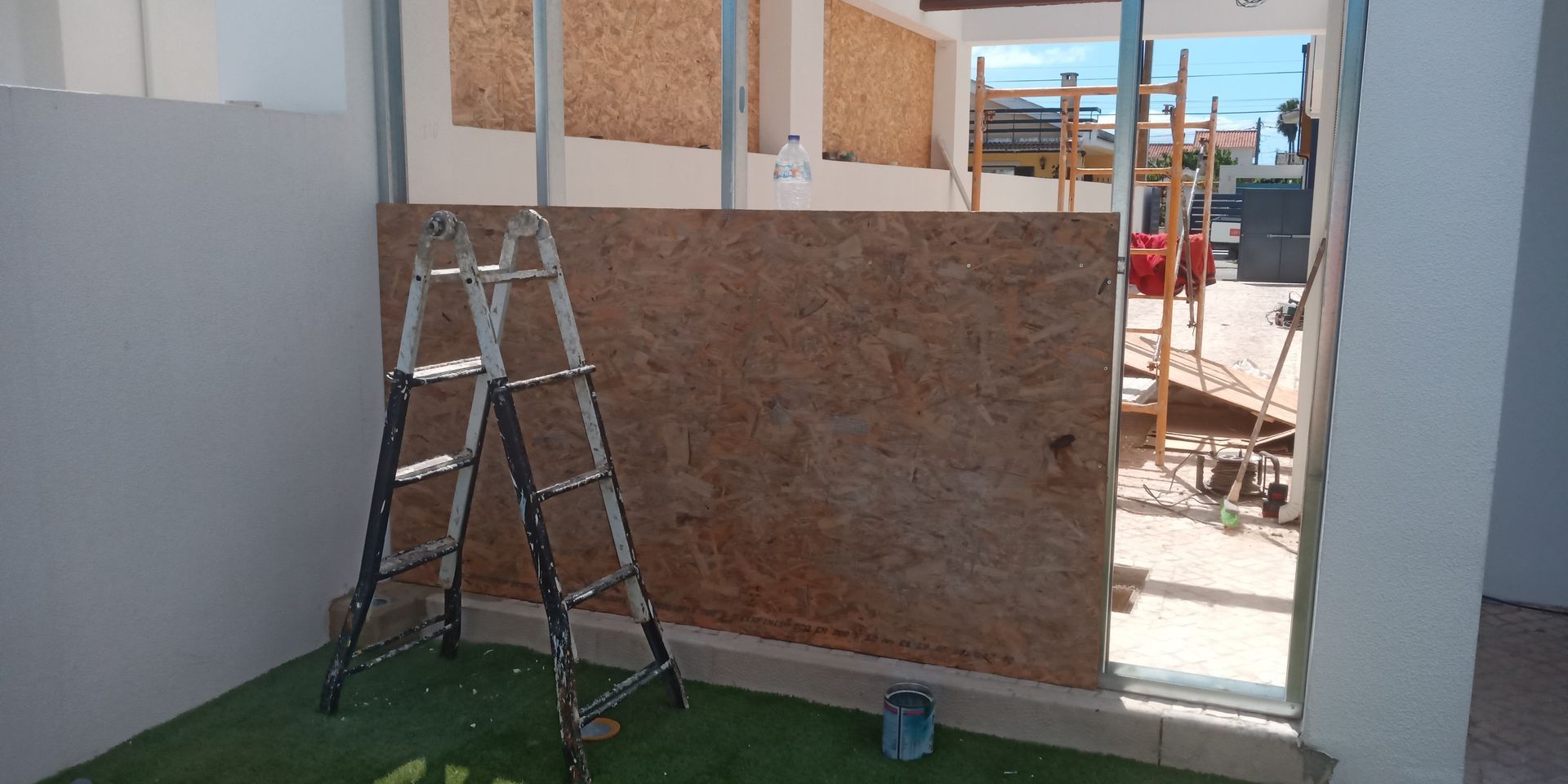 Canteiro de obras com escada. Painéis de madeira marrom estão fixados na estrutura. Grama artificial verde.