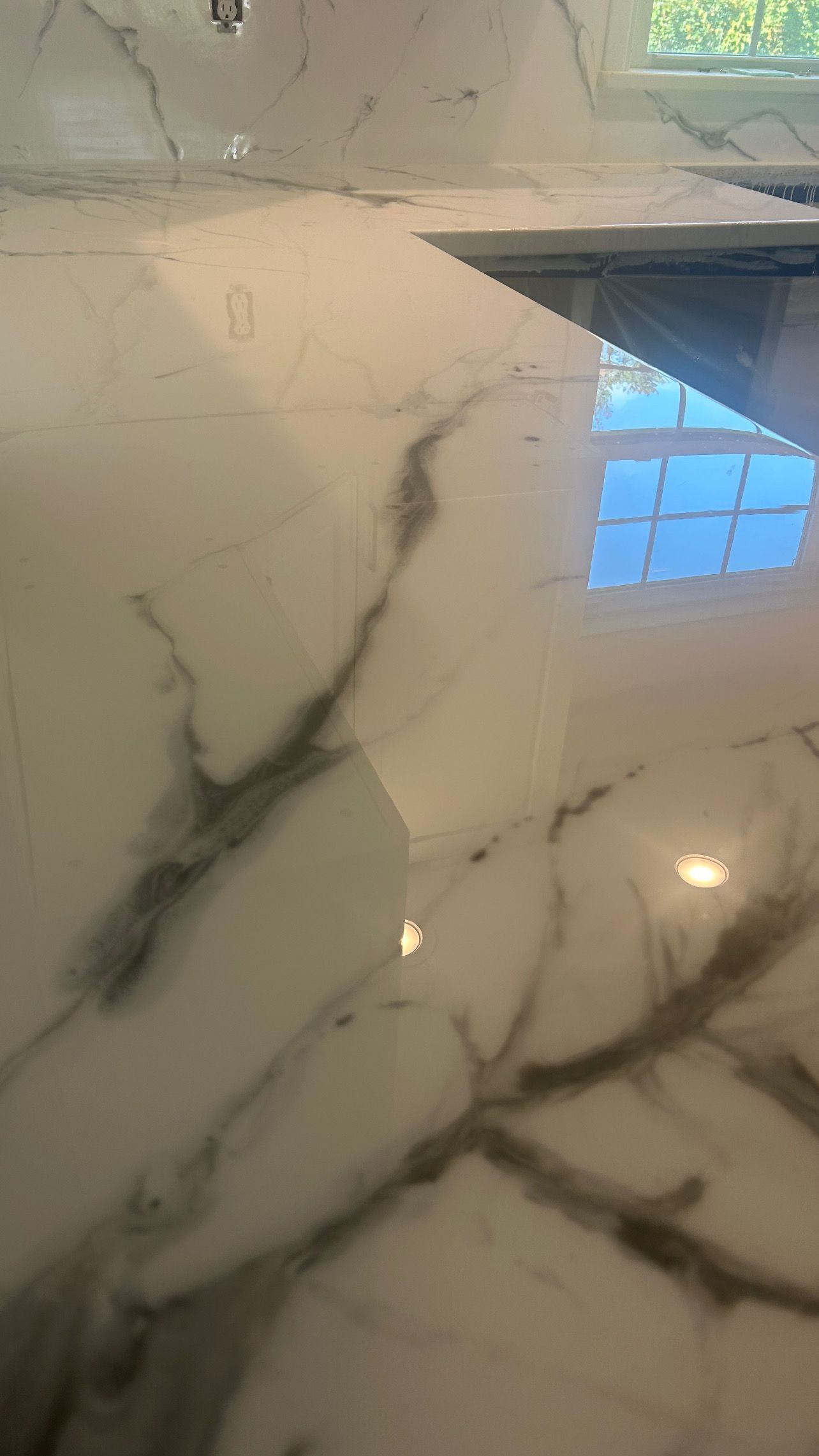 A high-angle view of a white kitchen countertop with grey veining, showing a reflection of a window.