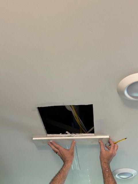 A person's hands holding a piece of white drywall up to a rectangular opening in a ceiling.