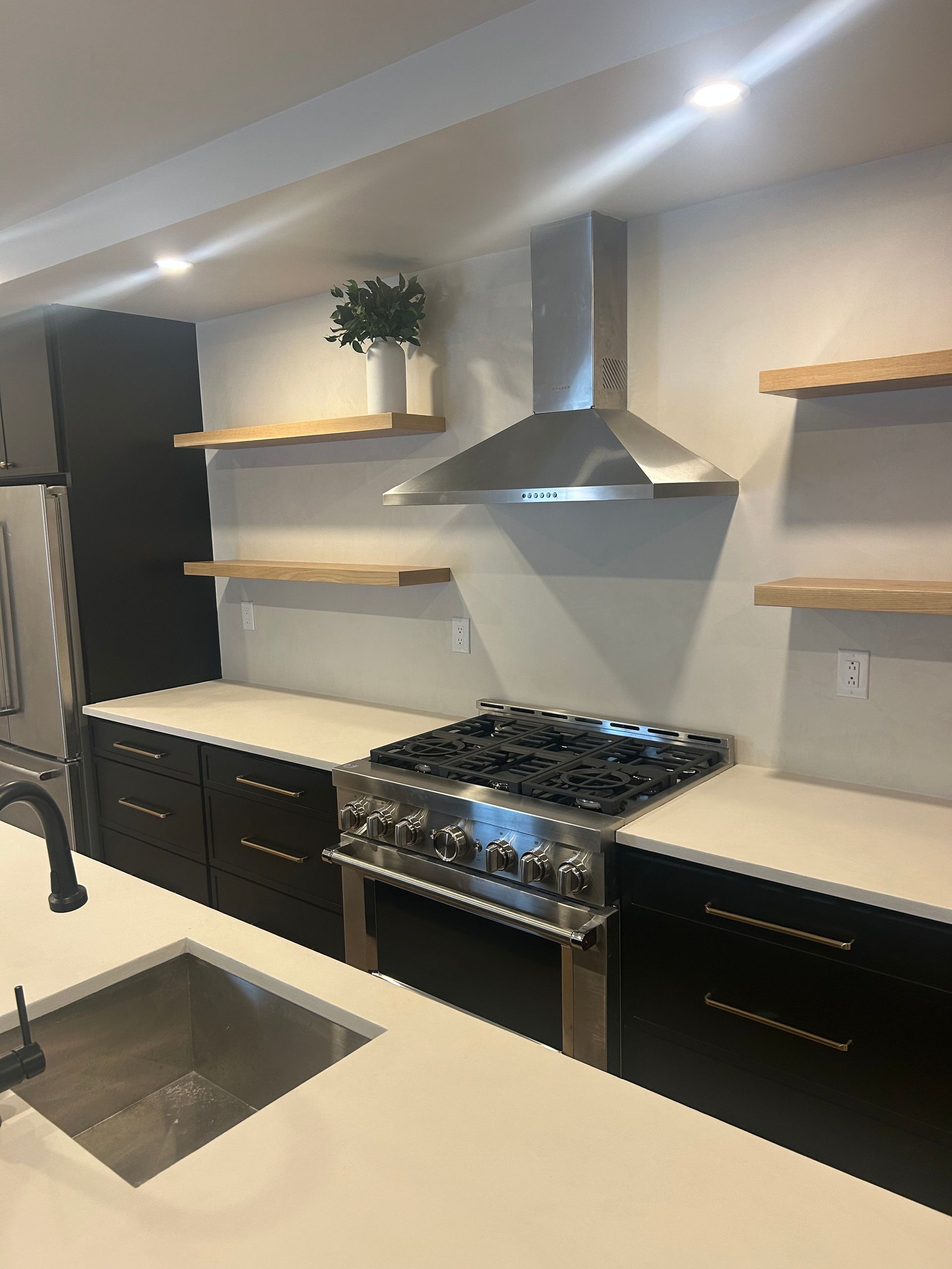 A modern kitchen featuring black cabinets, white countertops, a stainless steel gas range, and wood floating shelves.