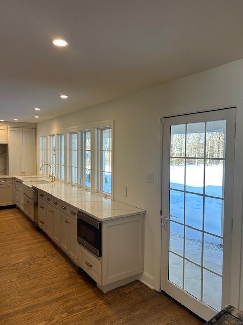 A modern kitchen with white cabinetry, quartz countertops, multiple windows, and a glass-paned door opening to snow.