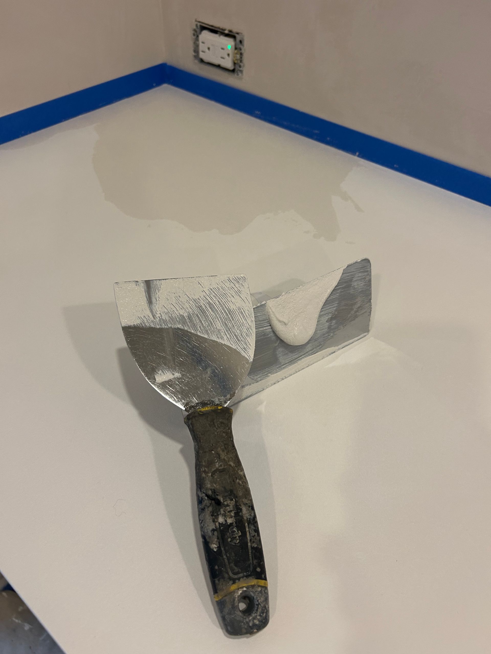 A putty knife with wet drywall compound resting on a primed wall surface with blue painter's tape along the corner.