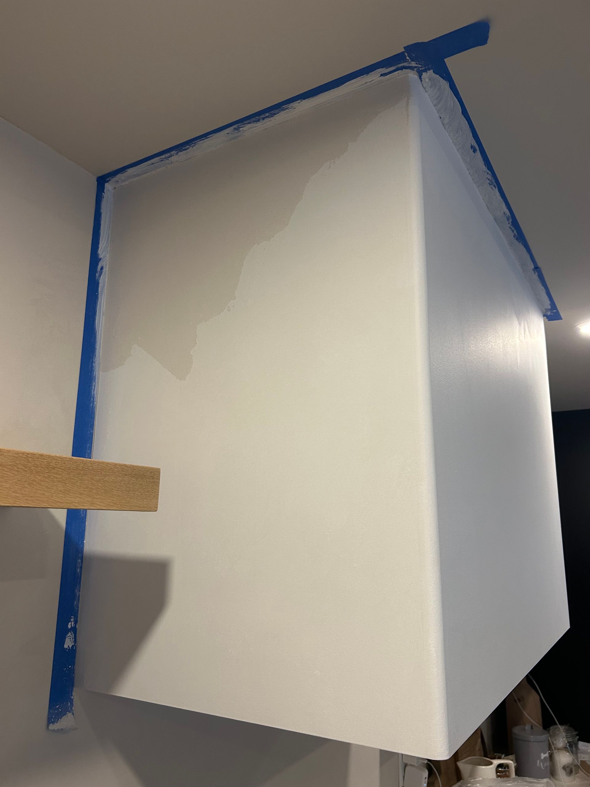 A white kitchen range hood under construction with blue painter's tape along the edges and a wooden shelf to the left.