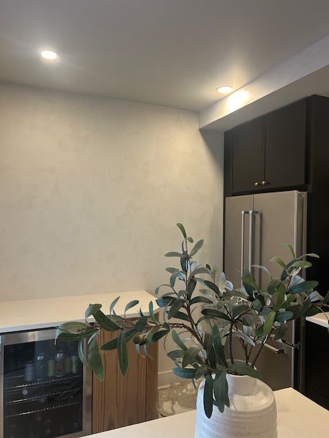 A white vase with green leafy branches sits on a counter in a modern kitchen with wooden cabinetry and a wine cooler.