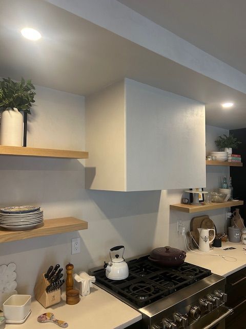 A modern kitchen with a white range hood, light wood floating shelves, a gas stove, and various kitchenware.