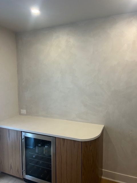 A built-in wood cabinet with a wine fridge beneath a white countertop, set against a wall with textured gray paint.