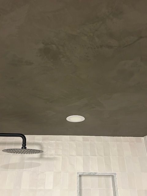 A ceiling with a recessed light and a shower head extending from the light-colored tiled wall below.