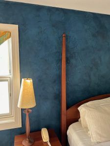 A gloved hand uses a wide metal putty knife to smooth plaster on a beige wall.