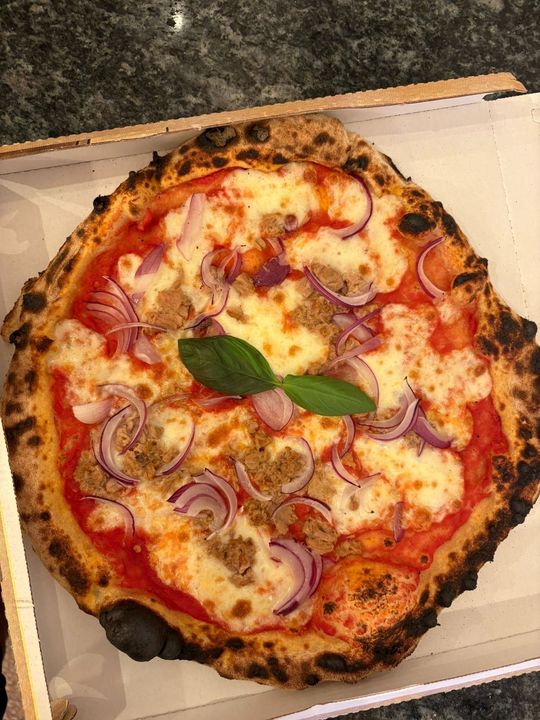 Pizza in scatola di cartone, con ingredienti che includono cipolla rossa, mozzarella e una foglia di basilico. La crosta è leggermente bruciacchiata.