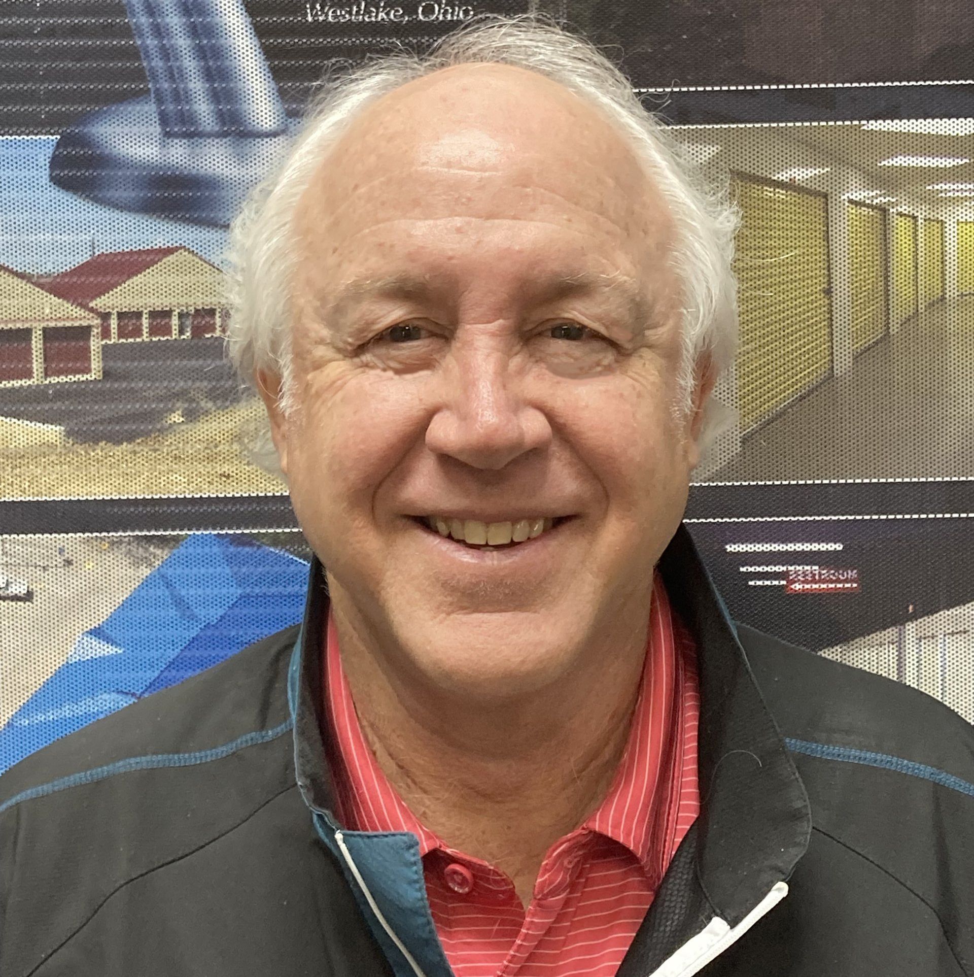 Smiling man in red shirt and black jacket. Background includes storage units.
