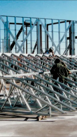 Workers installing metal trusses on a construction site. Steel frames and blue sky visible.