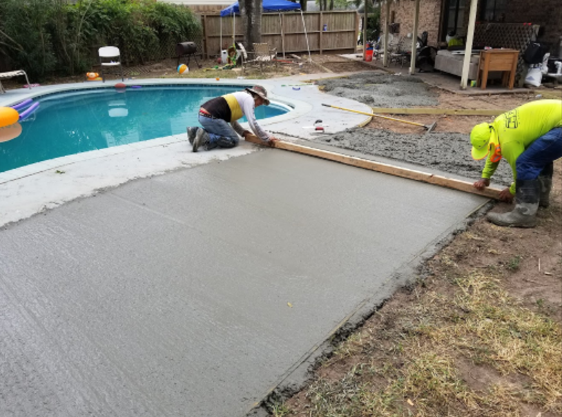 Two workers leveling wet concrete near a swimming pool. One kneels. Green, blue, and gray colors. Outdoors.