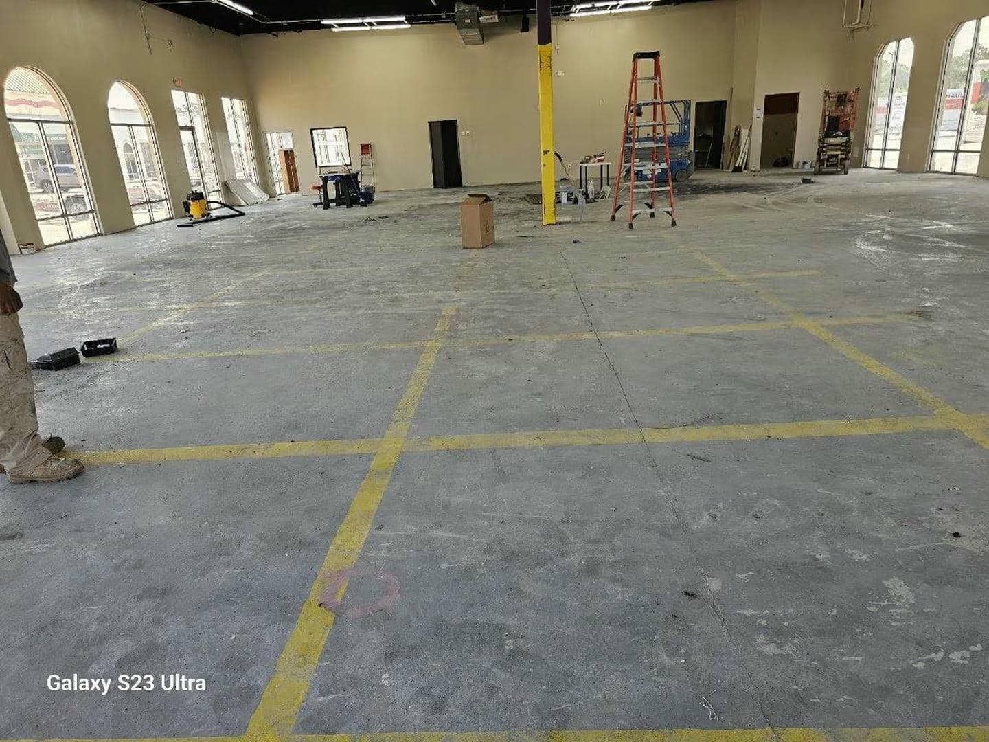 Large empty room undergoing renovations; gray floor with yellow lines, ladders, tools, and arched windows.