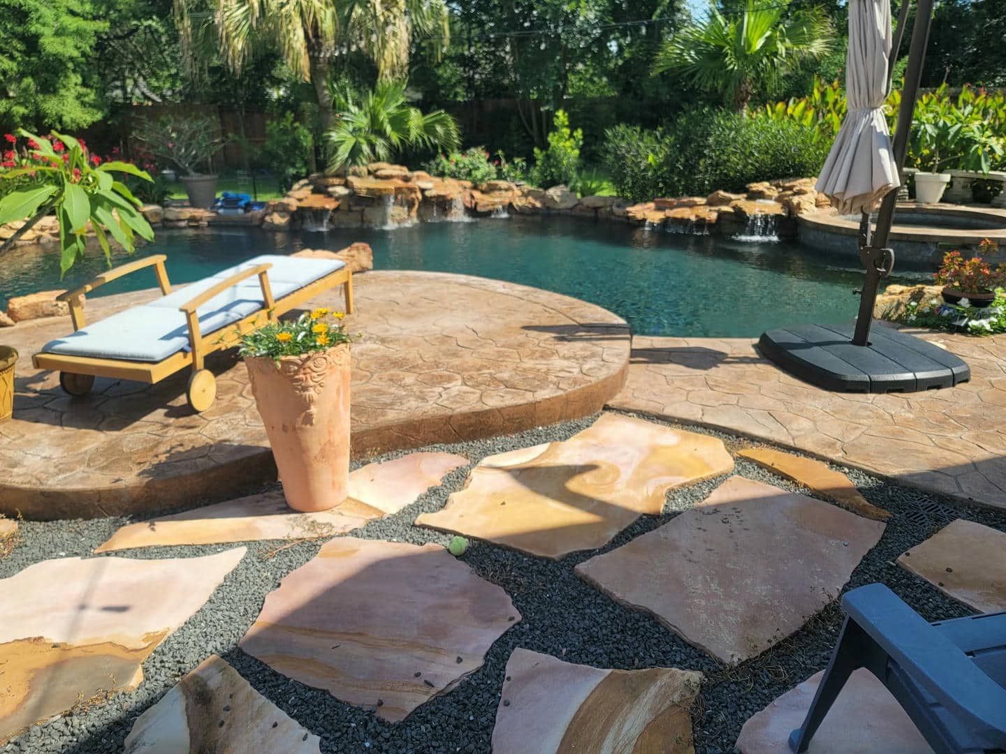 Poolside scene with natural stone patio, lounge chair, umbrella, and pool with small waterfall.