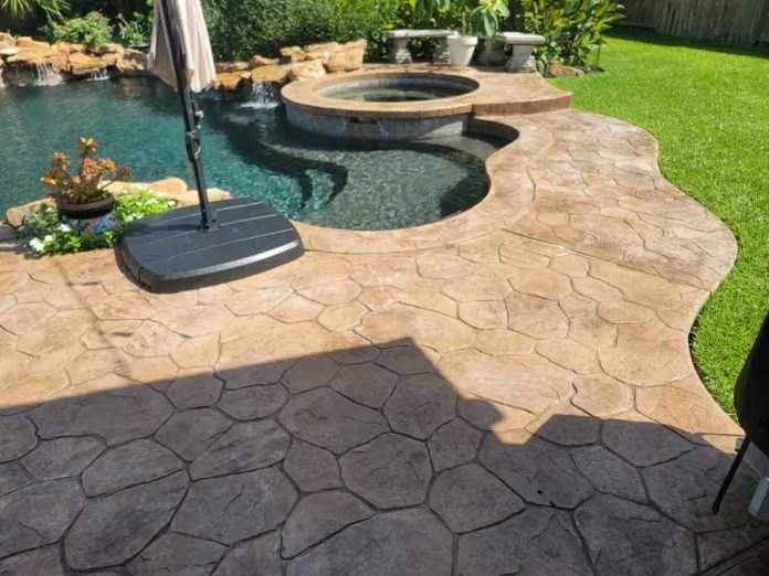 Pool and hot tub with stone-patterned concrete patio, umbrella, and green lawn.