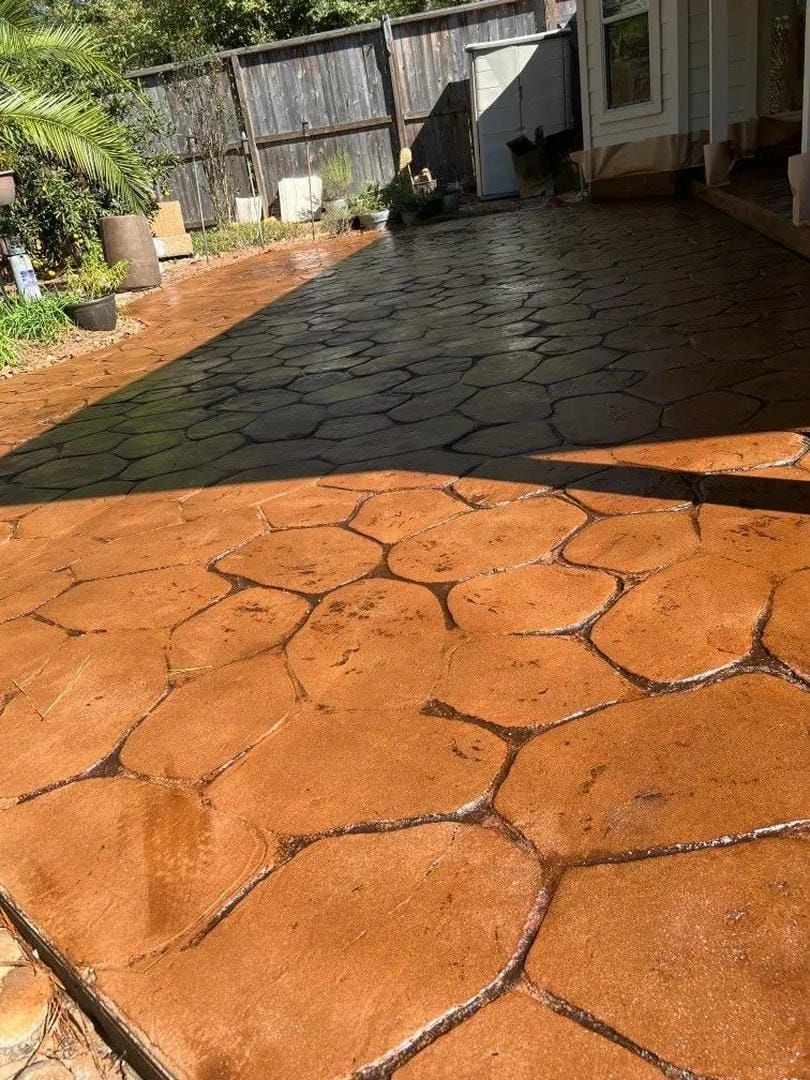 Stamped concrete patio, reddish-brown color, with irregular stone pattern. Sunlight reflects on the wet surface.