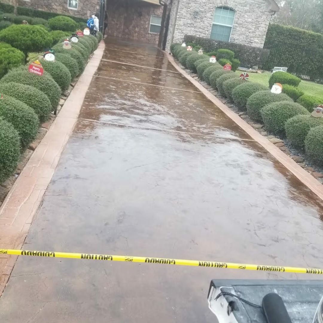 Wet, brown concrete path with yellow caution tape, lined with green bushes, next to a building.