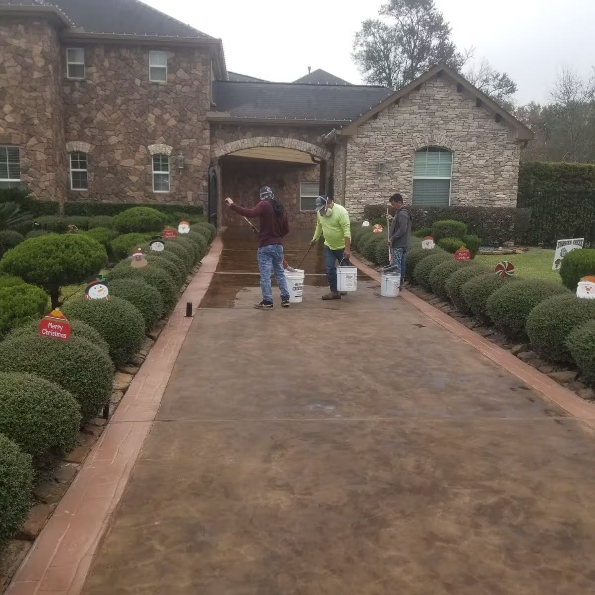 Three people applying sealant to a driveway in front of a stone house with manicured landscaping.