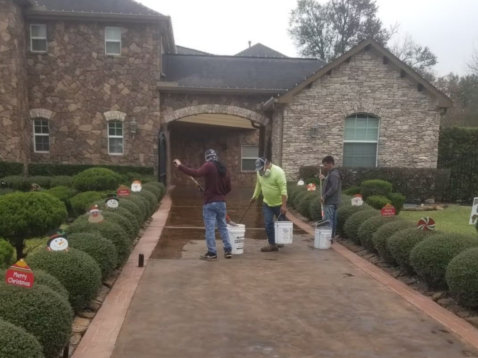 Three people painting a driveway in front of a stone house, wearing work attire.