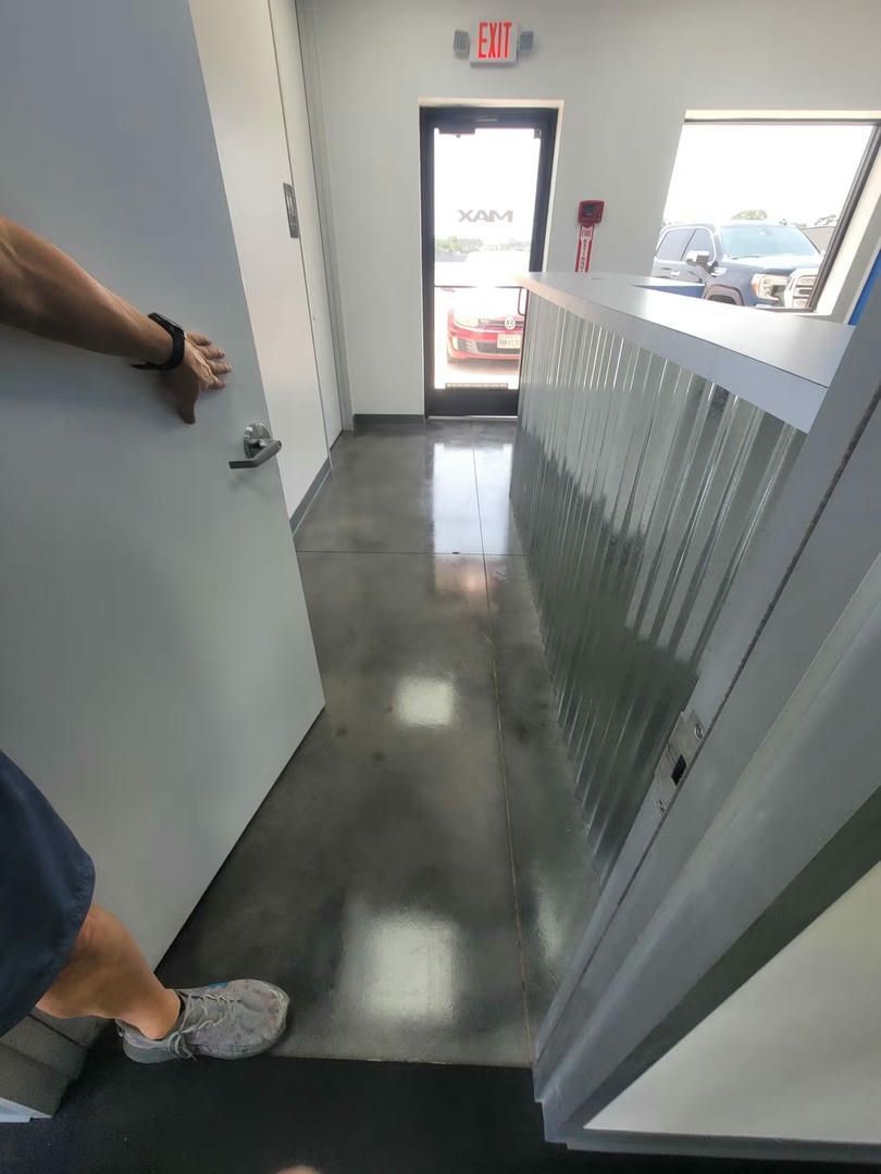 Person opening a door into a brightly lit office hallway with a concrete floor, exit sign, and a reception counter.