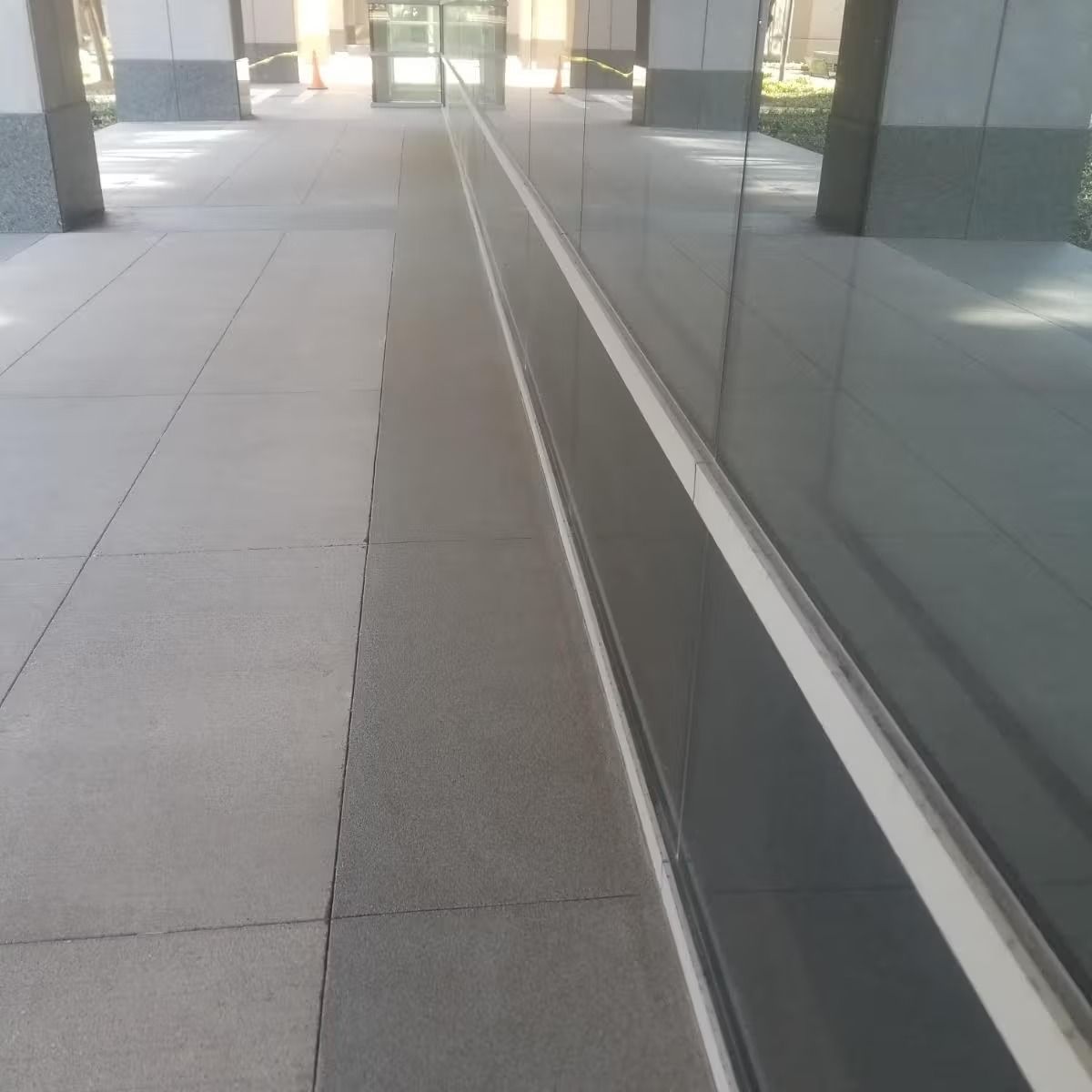 Exterior view of a building with a gray tiled walkway and a reflective glass wall. Sunlight creates shadows.