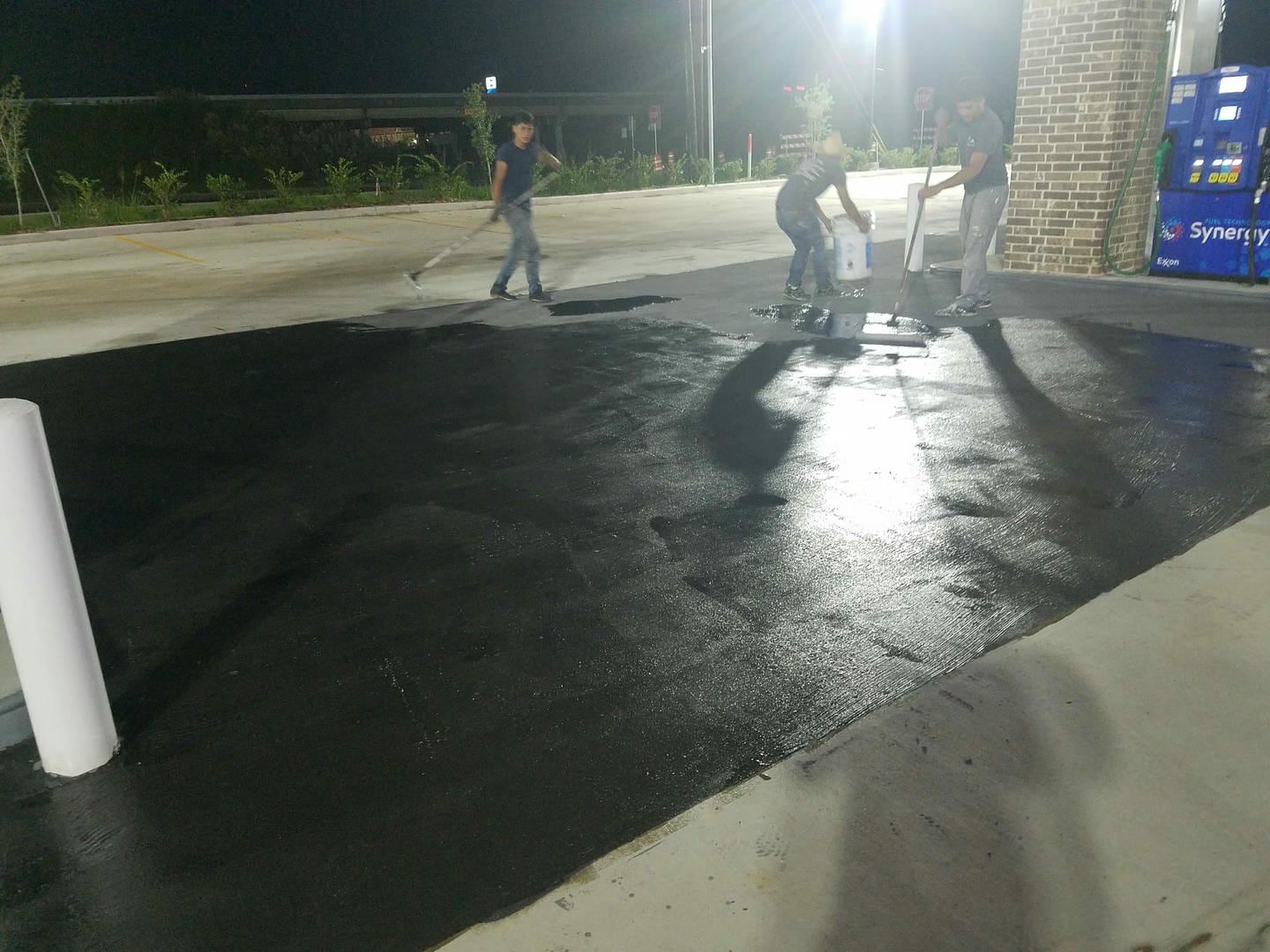 Workers paving asphalt at night, gas station setting.