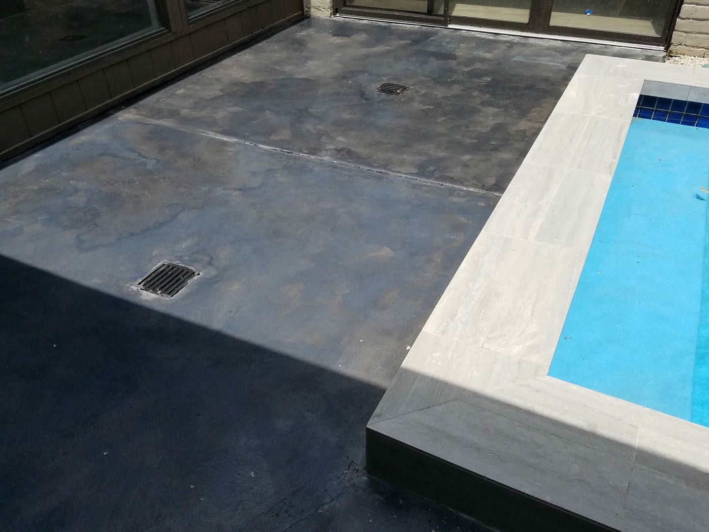 Concrete patio next to a pool with light blue water. Dark gray floor with drain. Bright sunlight.