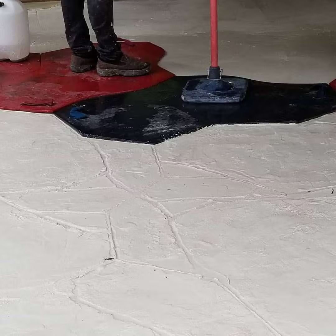 Person applying black epoxy coating to a concrete floor with a squeegee; white floor visible.