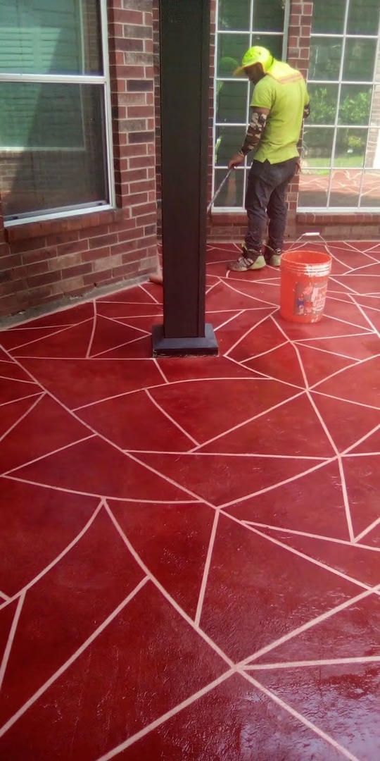 Man spraying sealant on red concrete patio with geometric white lines; window, brick wall.