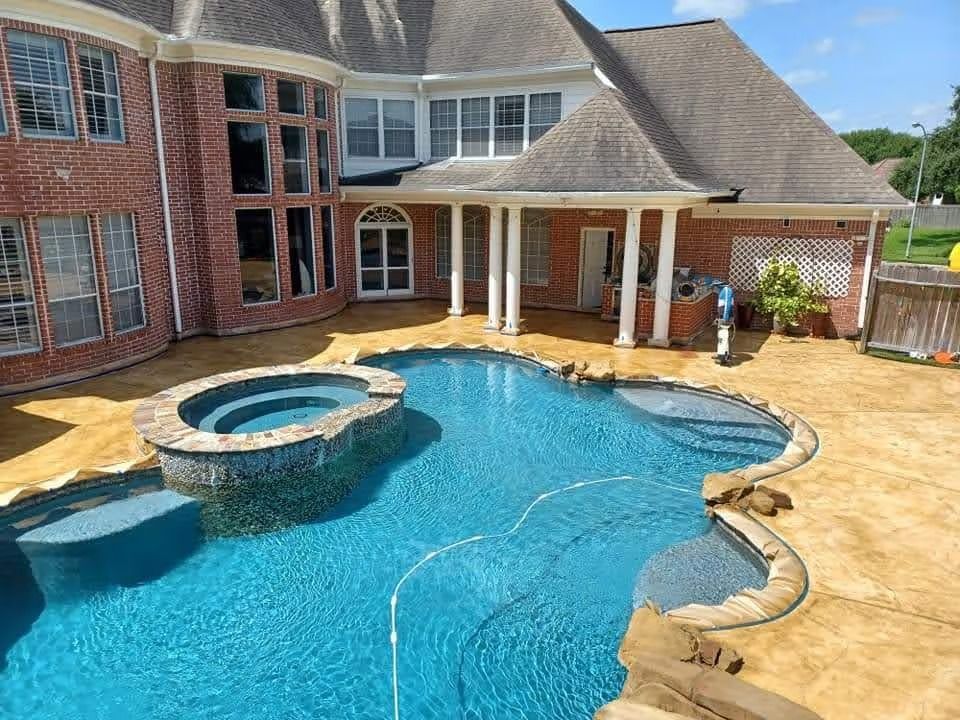 Backyard with a pool and spa next to a brick house. Sunny day with blue water and tan patio.
