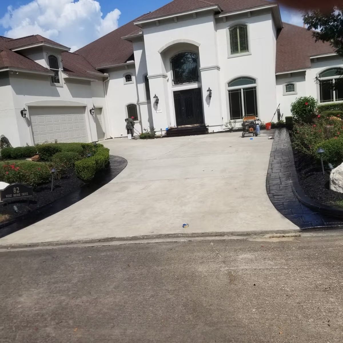 A large white house with a curved concrete driveway. Landscaping and a dark border line the driveway.