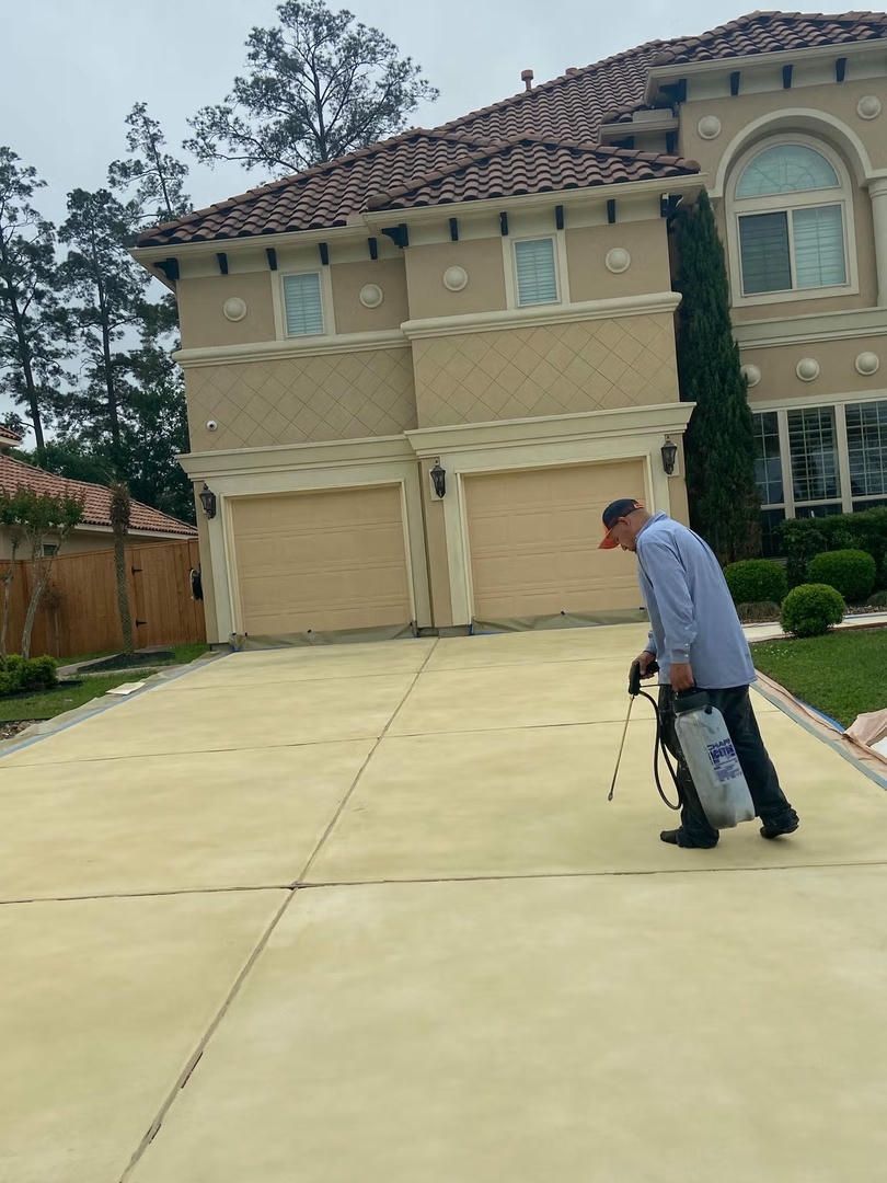 Man spraying a driveway in front of a large house with a tile roof and two garage doors.