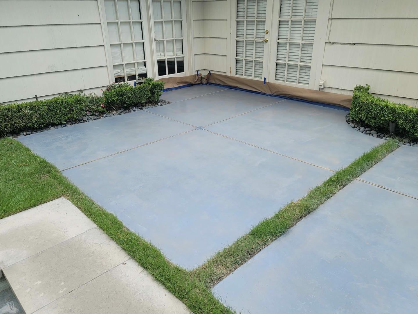 Blue painted patio with grass borders, bushes, and white siding.