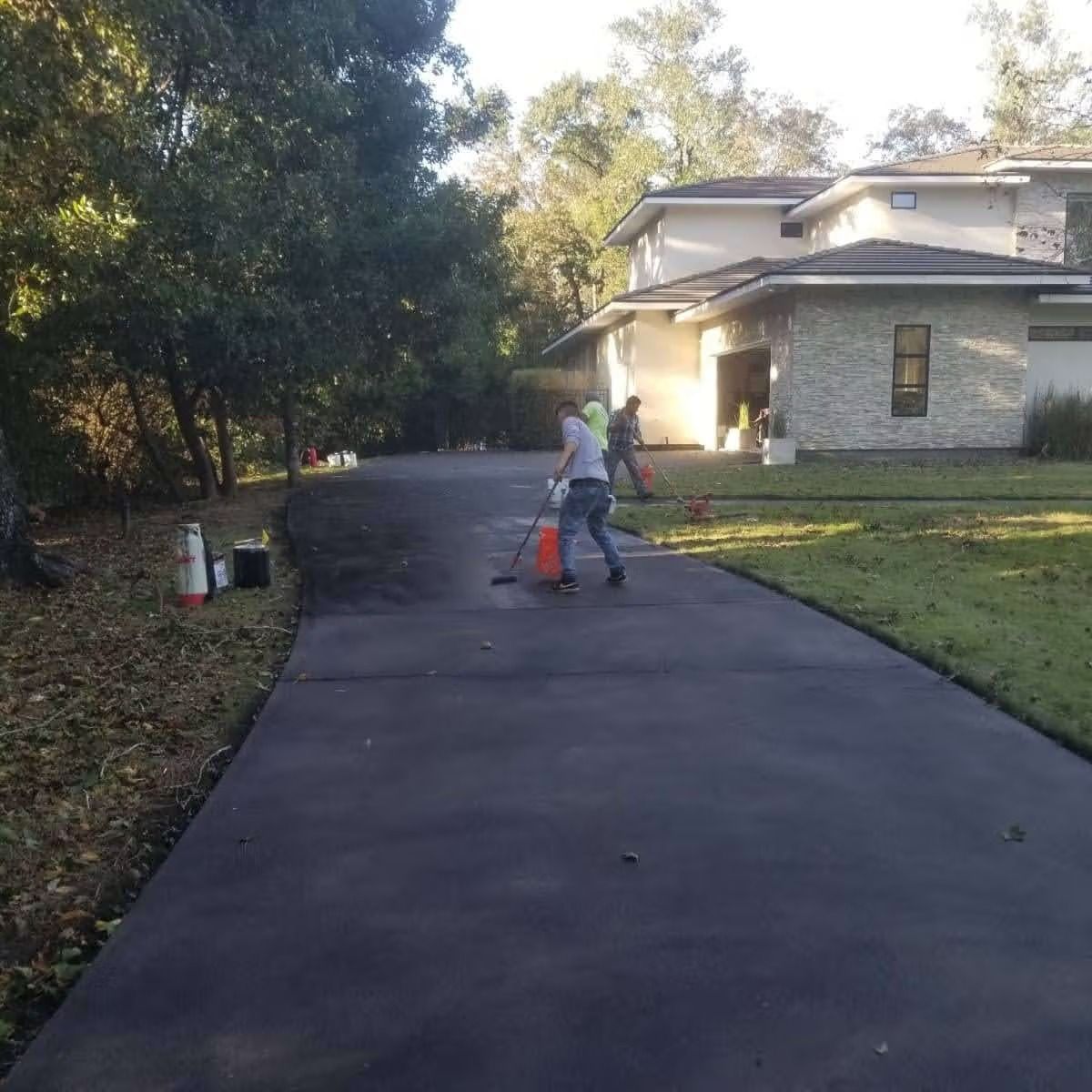 Workers seal coating a long asphalt driveway in front of a modern house.
