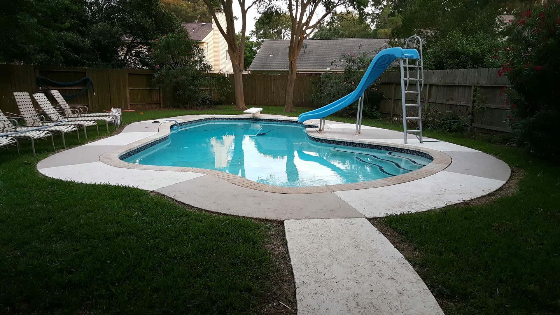 Backyard pool with a blue slide, surrounded by a concrete deck and green grass.