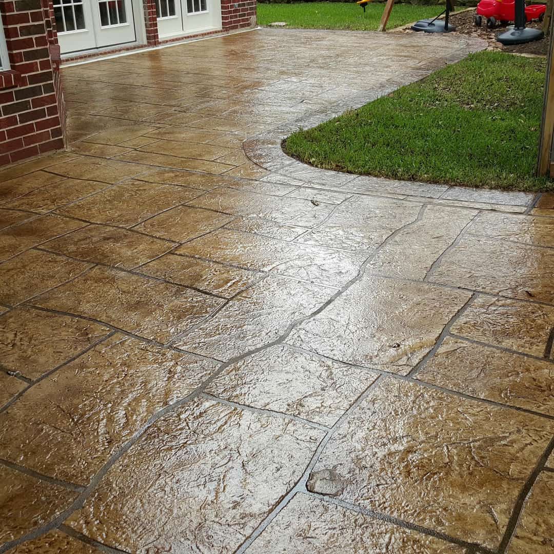 Stamped concrete patio with a wet, brown, stone-like appearance; adjacent to green grass and brick wall.