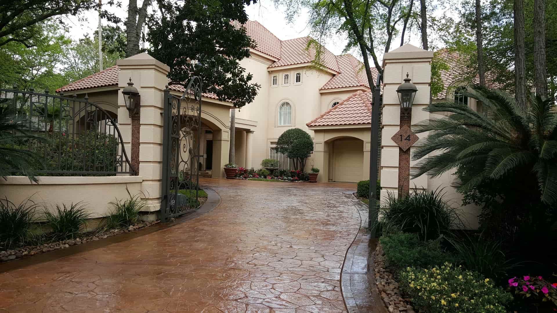 Beige stucco house with red tile roof, long driveway, and wrought iron gate.