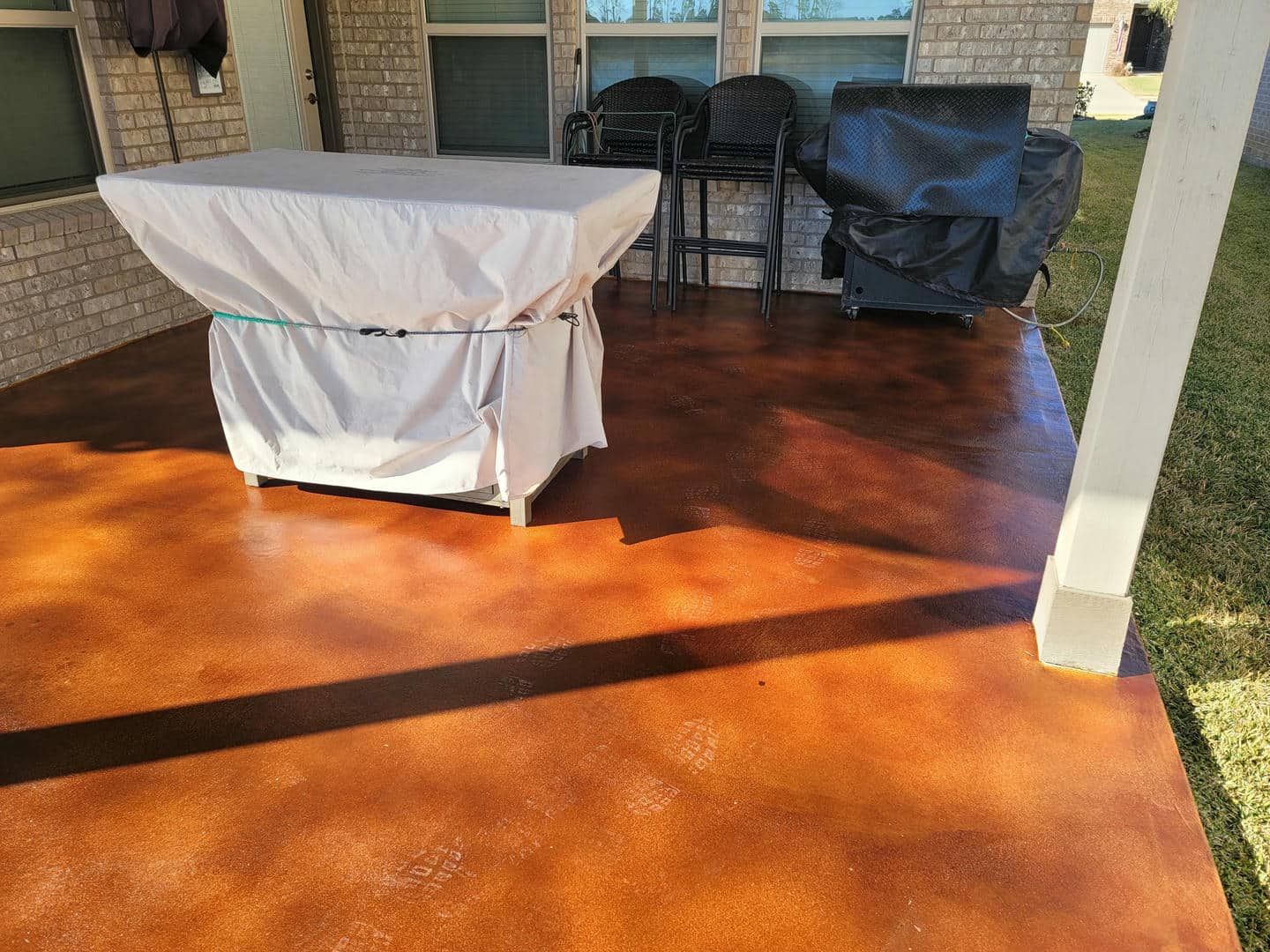 Patio with stained concrete, covered table, chairs, and grill. Brown tones with sunlight.