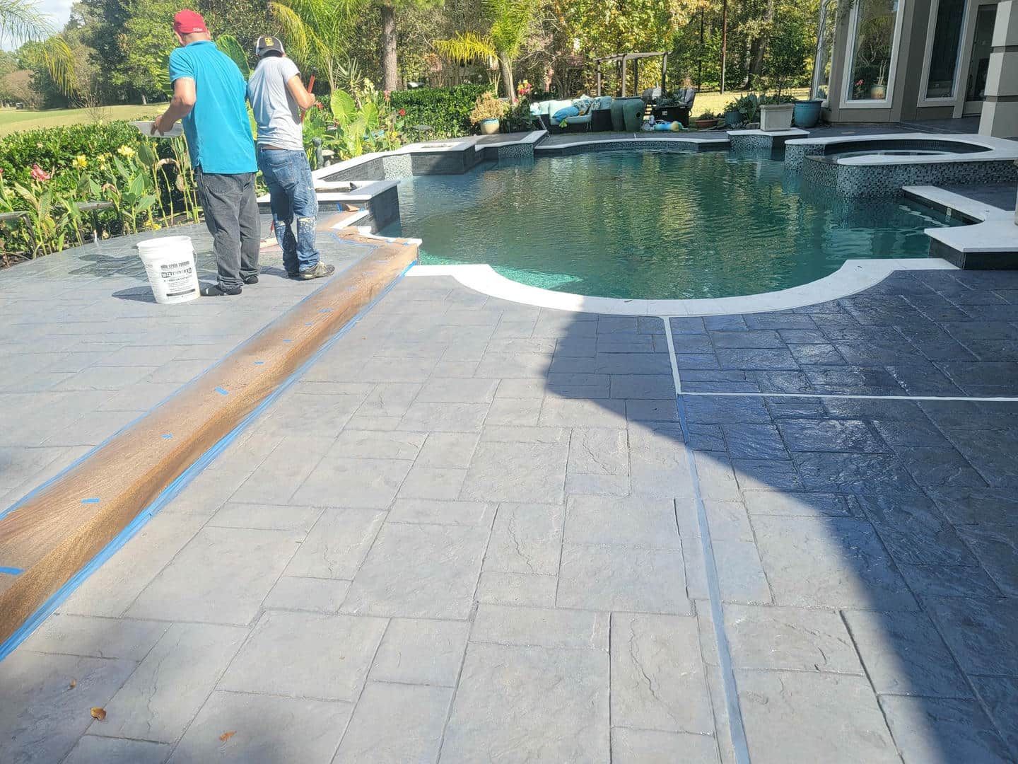 Two workers staining a concrete patio surrounding a swimming pool.