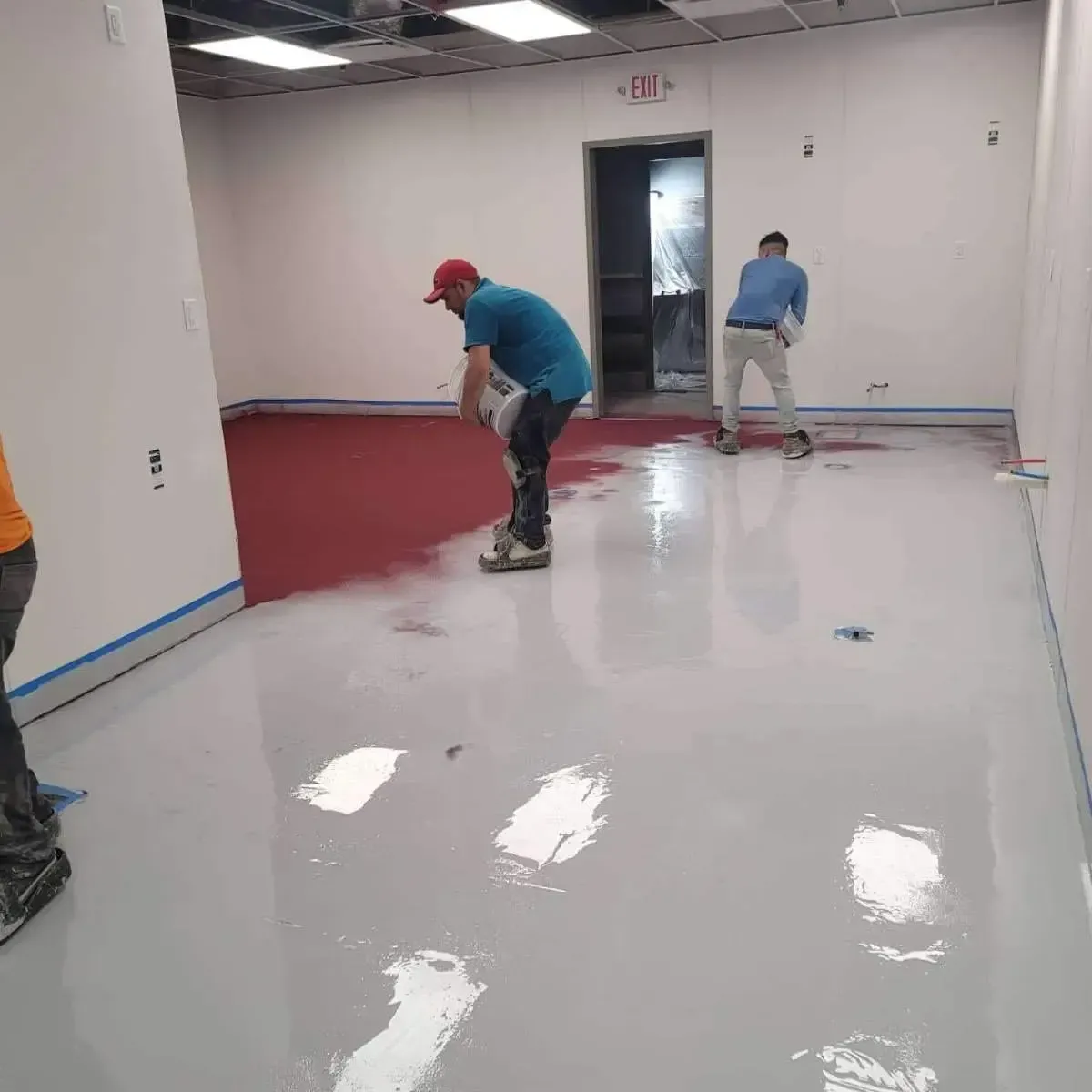 Workers applying red and gray epoxy flooring in a room with white walls.