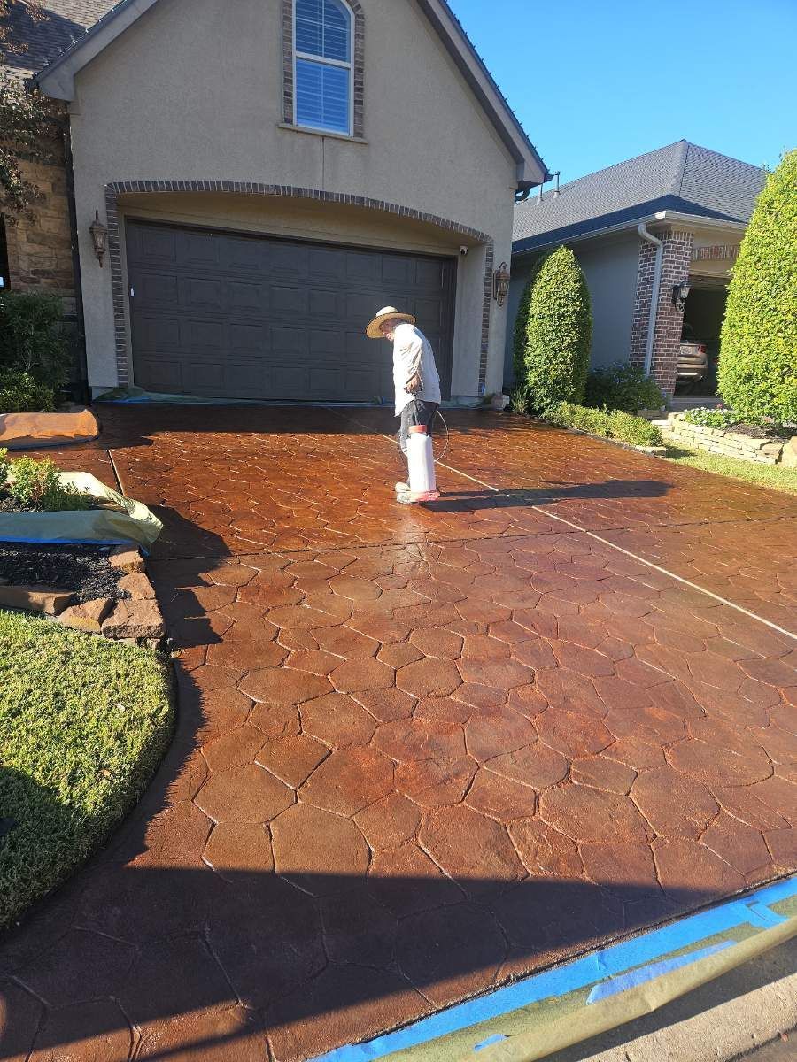 Person sealing a stamped concrete driveway, two-car garage in the background. Brown and reddish tones.