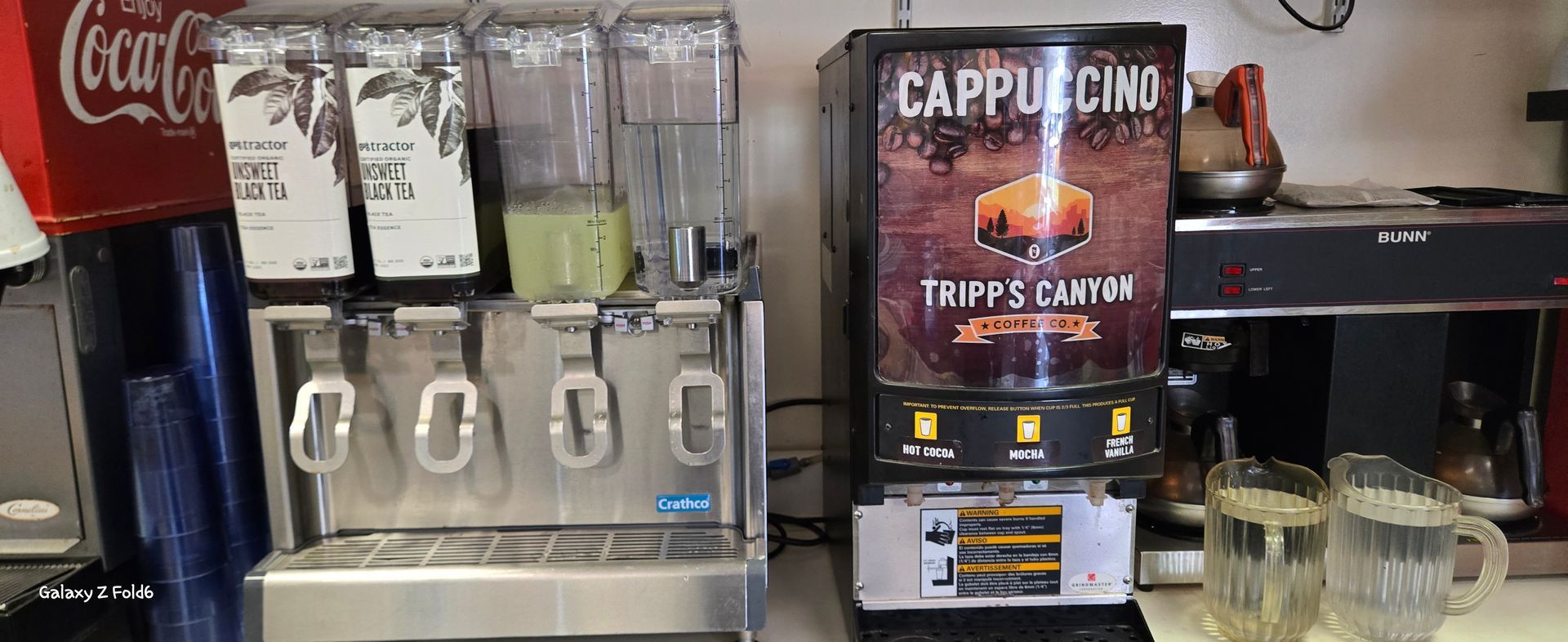 A cappuccino machine is sitting on a counter next to a coca cola machine.