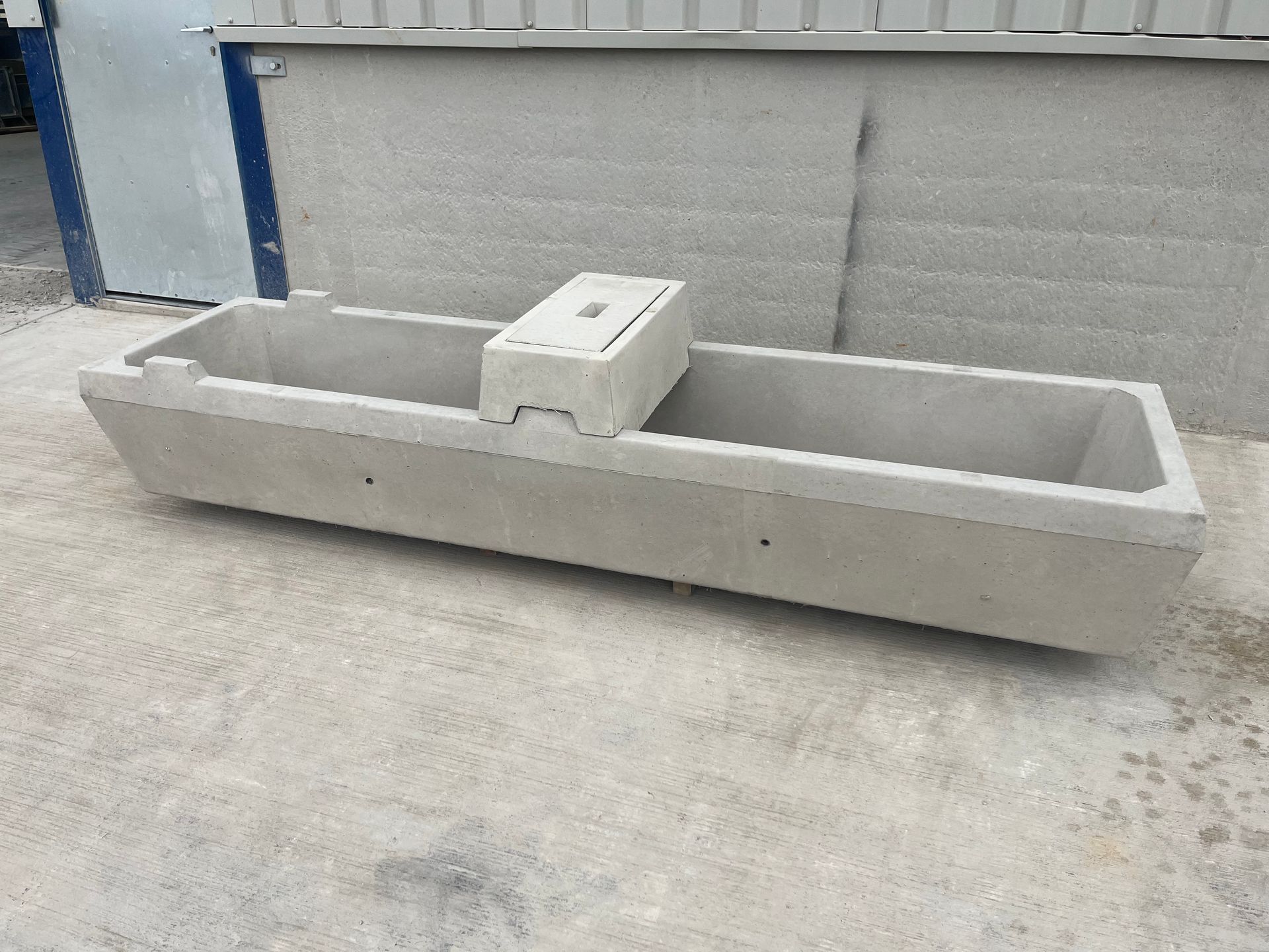 A long, grey concrete livestock watering trough with a central service compartment, sitting on a concrete surface.