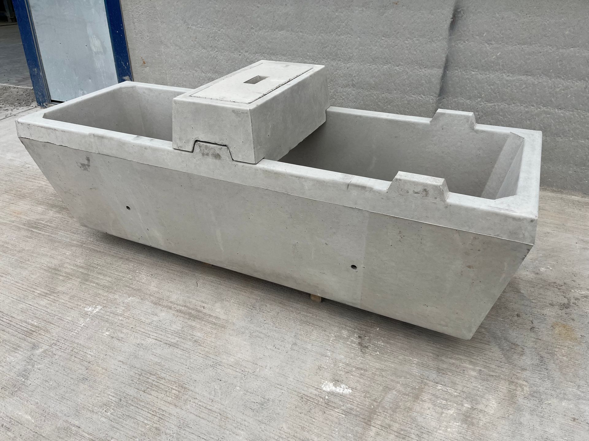 A rectangular concrete trough or livestock waterer with a central access cover, sitting on a concrete surface.